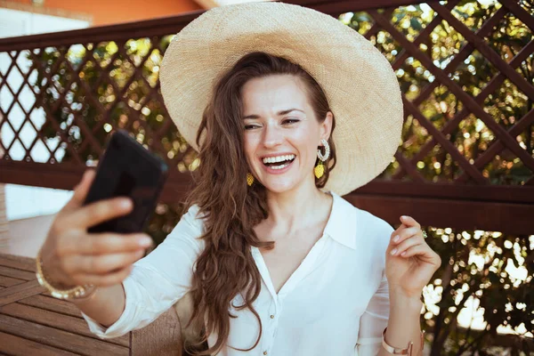 幸せな現代的な中年主婦の白いシャツに帽子を持っていますウェブカメラ会議でテラスでスマートフォン — ストック写真