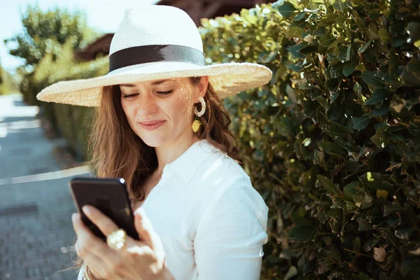 緑の壁の近くの屋外でスマートフォンのアプリケーションを使用して帽子を持つ白いシャツの現代女性 — ストック写真