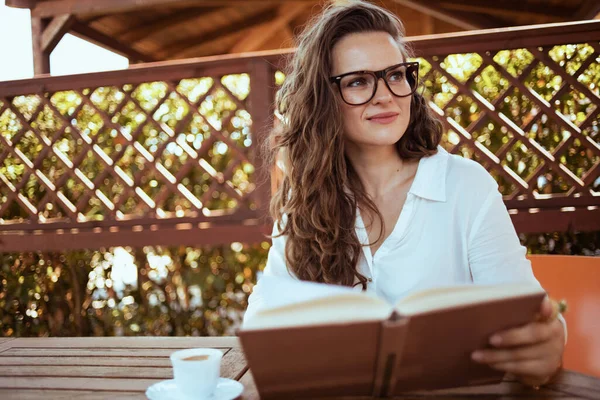 40歳の優雅な白いシャツにコーヒーと本と眼鏡をかけた女性がテラスのテーブルに座って — ストック写真