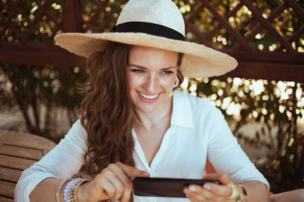 40歳の白いシャツを着た女性を笑顔にし ゲストハウスホテルのパティオでスマートフォンを使ってテキストメッセージを送る — ストック写真