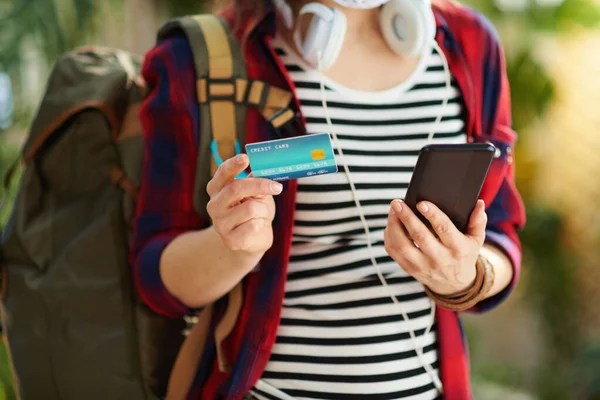 Travel during covid-19 pandemic. Closeup on trendy traveller woman with white medical mask, backpack, credit card and smartphone booking tickets online in the modern house in sunny day.