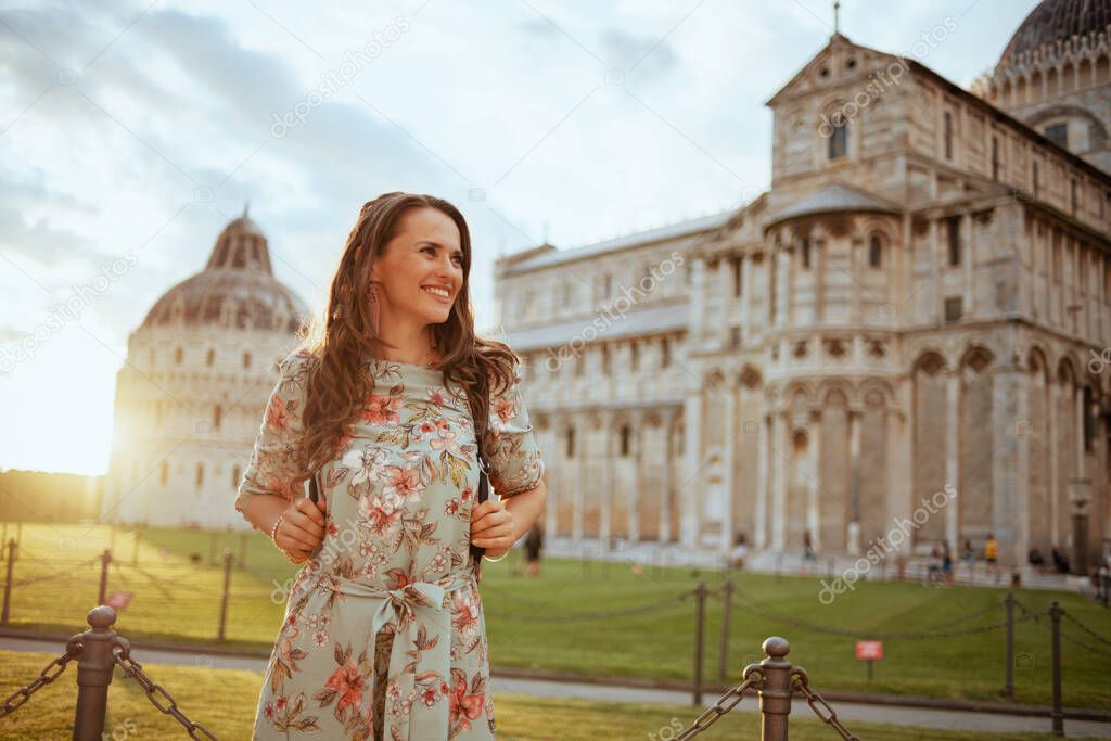 smiling young woman in floral dress with backpack having walking tour in piazza dei miracoli in Pisa, Italy.