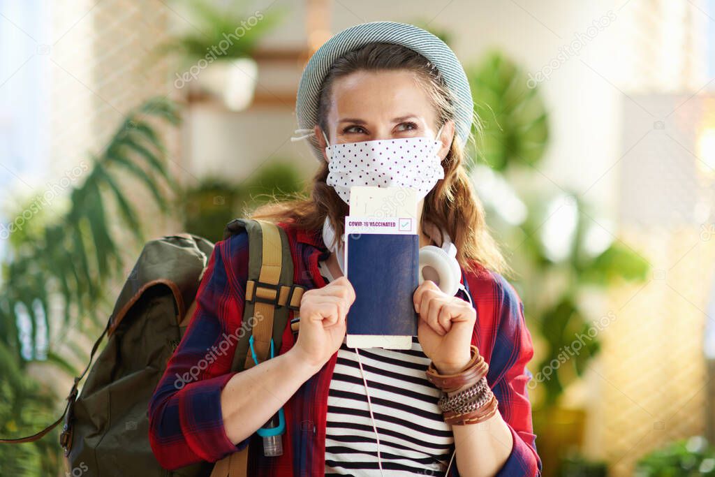 Travel during covid-19 pandemic. pensive trendy solo traveller woman with covid passport, white medical mask and backpack in the modern living room in sunny day.