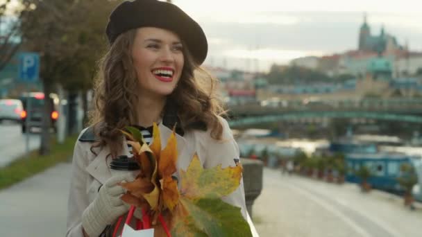 城市街道上 身穿米黄色风衣 头戴黑色贝雷帽 头戴购物袋 一杯热巧克力和秋天黄叶的中年妇女面带笑容 — 图库视频影像