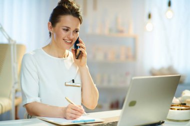 Bilgisayarlı gülümseyen kadın çalışan modern güzellik stüdyosunda akıllı telefondan konuşuyor..