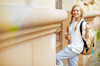 Çalışma kitabı, dezenfektan ve sırt çantası olan beyaz tişörtlü, şık bir çocuk şehirde el sallıyor ve okula gidiyor..