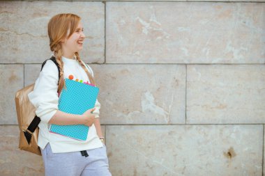 Beyaz tişörtlü, çalışma kitabı ve sırt çantasıyla şehrin dışında duvara yaslanmış sırıtan modacı kız..