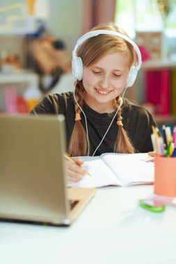 Gri tişörtlü, laptopu, kulaklığı ve çalışma kitabı olan mutlu modern öğrenci güneşli bir günde evde online eğitim görüyor..