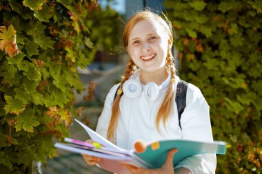 Şehirdeki yeşil okulun yakınlarında iş kitabı, sırt çantası ve kulaklığı olan beyaz kazaklı mutlu bir çocuğun portresi..