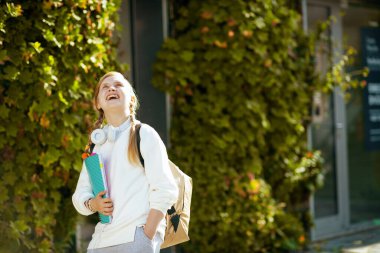 Çalışma kitabı, sırt çantası ve kulaklığı olan beyaz tişörtlü mutlu, modern bir kız şehirdeki yeşil okulun yakınına bakıyor..