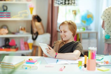 Gri tişörtlü, tabletli ve çalışma kitabıyla gülümseyen modern çocuk güneşli bir günde evde ders çalışıyor..