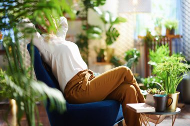 Yeşil Ev. Modern evdeki mavi koltukta oturan yeşil pantolonlu, gri bluzlu, uzun dalgalı saçlı modern ev hanımı..