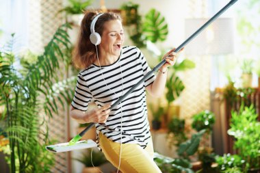 Sırıtan orta yaşlı, çizgili tişörtlü, paspas ve kulaklıklı bir kadın güneşli bir günde modern oturma odasında temizlik yapıyor..