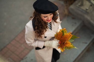 Merhaba Eylül. Sonbaharda şehir sokaklarında sonbahar sarısı yaprakları olan bej renkli trençkotlu ve siyah bereli gülümseyen trend kadın manzarası..