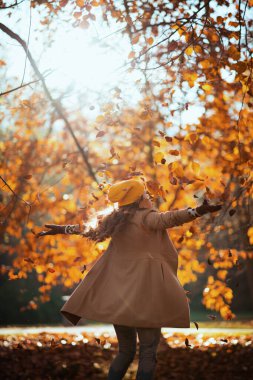 Merhaba sonbahar. Bej ceketli, turuncu şapkalı güler yüzlü bir kadın sonbaharda şehir parkında neşe saçıyor..