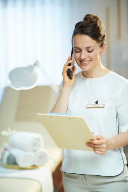 Kırk yıllık gülümseyen kadın çalışan bir pano ile modern güzellik salonunda akıllı bir telefonla konuşuyor..