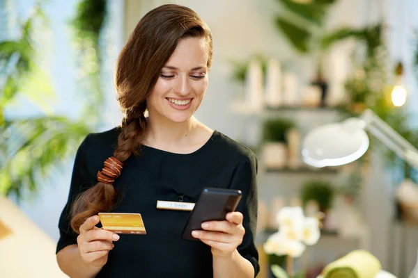 スマートフォンとクレジットカードで幸せな女性労働者は 現代の美容室で電子商取引サイトでオンラインショッピングを作る — ストック写真