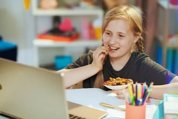 幸せな現代の女の子でグレーシャツでノートパソコンの距離学習と食べる健康スナックに家で晴れた日 — ストック写真