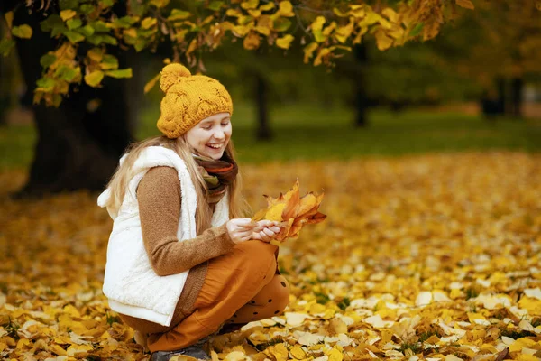穿着棕色毛衣 头戴橙色帽子 面带微笑的小孩在城市公园的室外采摘秋天的树叶 — 图库照片
