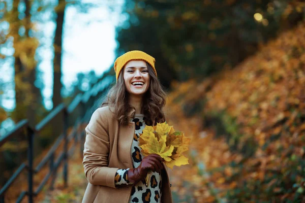 城市公园里穿着棕色外套 头戴黄色帽子 头戴秋天黄叶的中年妇女在户外微笑着 — 图库照片