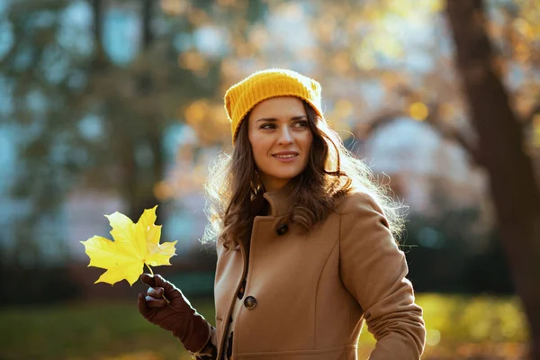 ベージュコートの幸せな若い女性と秋に街の屋外で黄色の葉とオレンジの帽子 — ストック写真