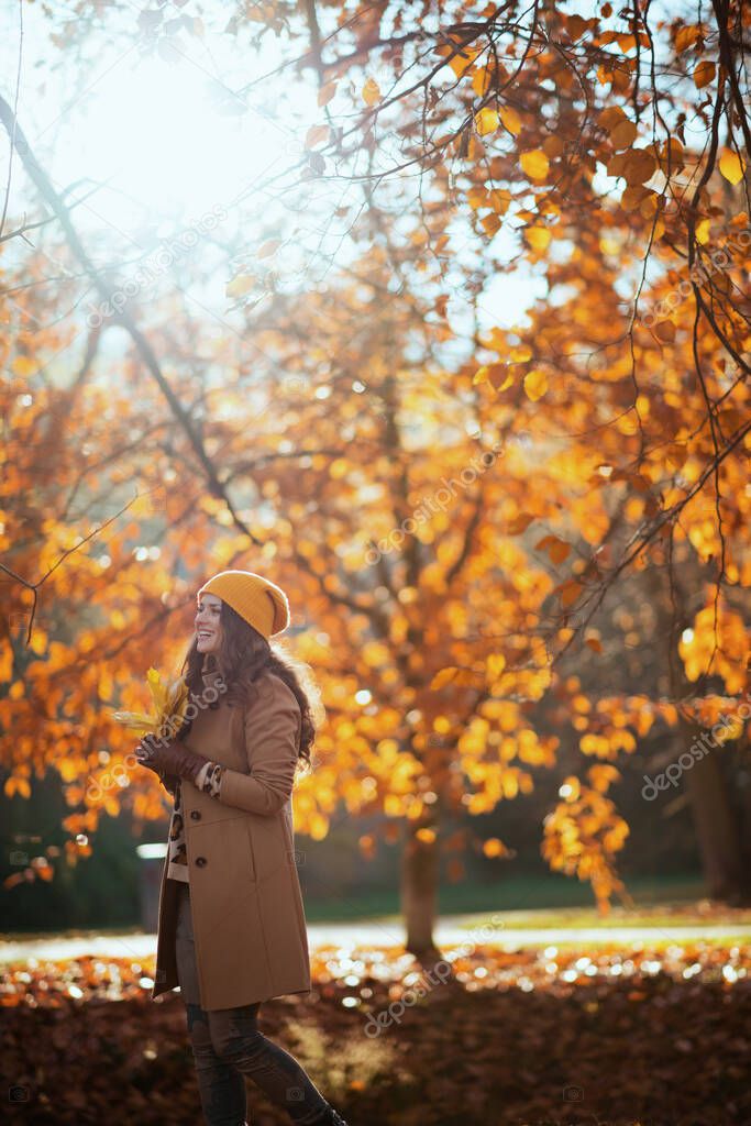 Hello november. happy young woman in beige coat and orange hat with autumn yellow leaves outdoors on the city park in autumn.