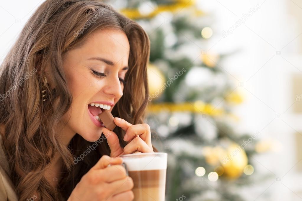 Young woman eating candy with latte macchiato near christmas tre