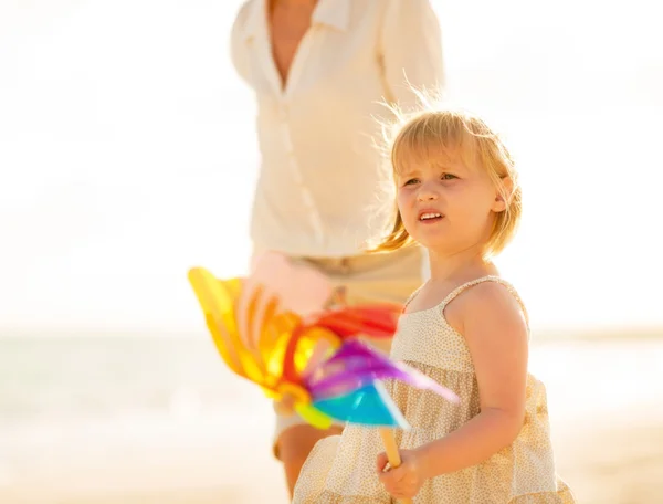 Mother and baby girl with toy windmill walking on the beach in t — Stock Photo, Image