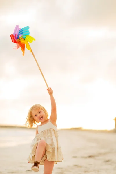 Happy baby girl playing with colorful windmill toy on the beach — Stock Photo, Image