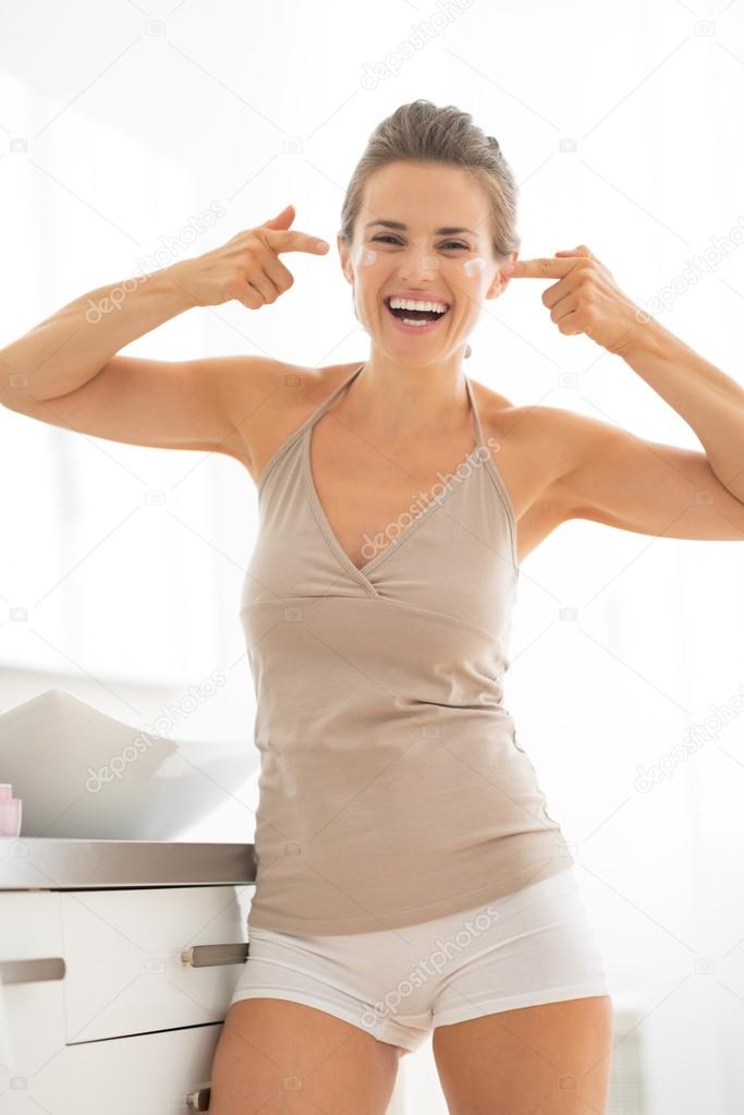 Smiling young woman applying cream in bathroom