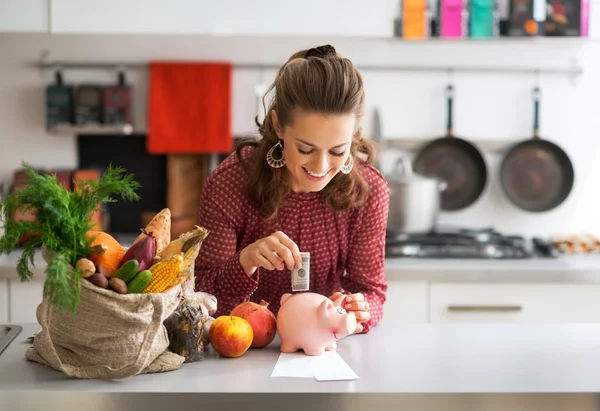 Retrato de la joven ama de casa feliz poniendo dinero en alcancía — Foto de Stock