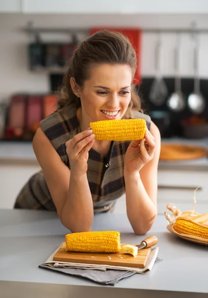 Retrato de feliz ama de casa joven comiendo maíz hervido en la cocina — Foto de Stock