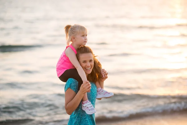 Ευτυχισμένο μωρό κορίτσι που κάθεται στους ώμους της μητέρας στην παραλία στο e — Φωτογραφία Αρχείου