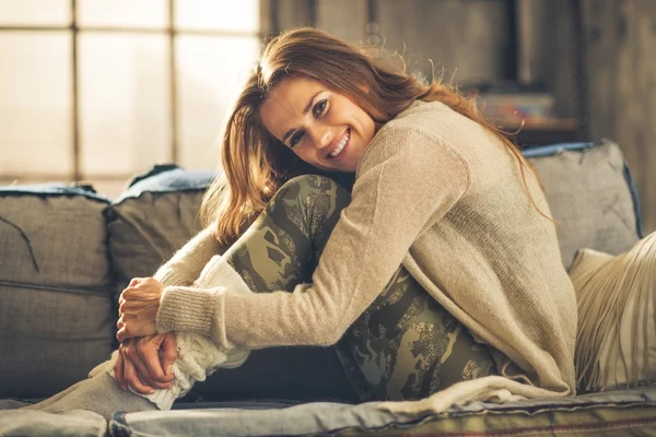 Uśmiechnięta kobieta na kanapie w poddaszu, tulenie kolana — Zdjęcie stockowe