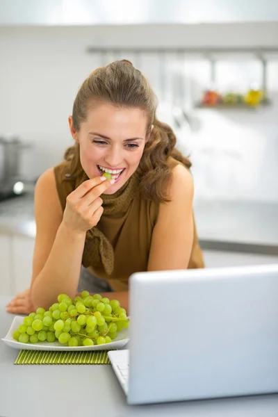 Happy young woman eating grape in kitchen and using laptop — Stock Photo, Image