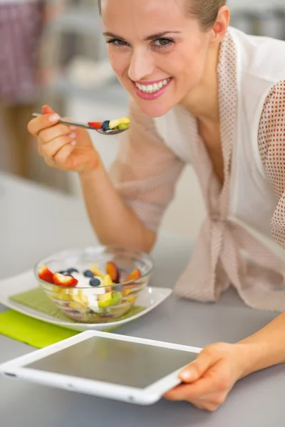 Glückliche junge Frau isst Obstsalat und benutzt Tablet-PC — Stockfoto