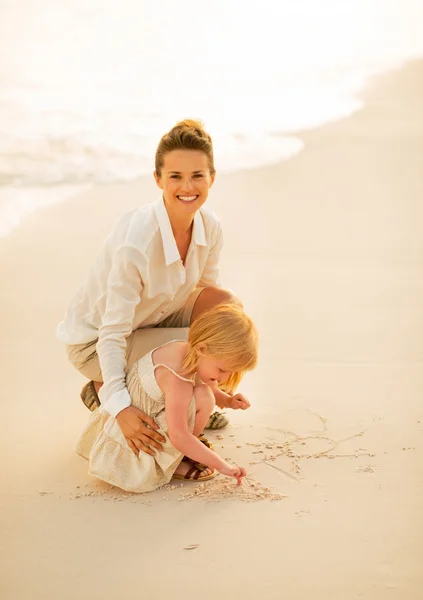 Glückliche Mutter und kleines Mädchen zeichnen auf Sand am Strand am eveni — Stockfoto