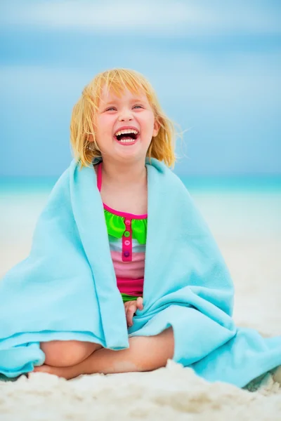 Portrait of laughing baby girl wrapped in towel sitting on beach — Stock Photo, Image