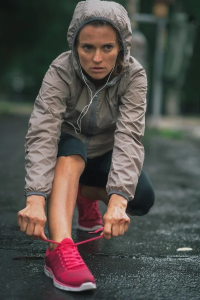Fitness young woman tying shoelaces outdoors in rainy city — Stock Photo, Image