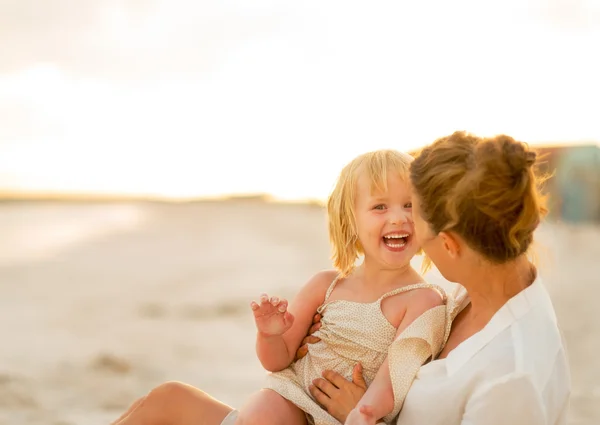 Moeder en baby meisje, zittend op het strand in de avond lachen — Stockfoto