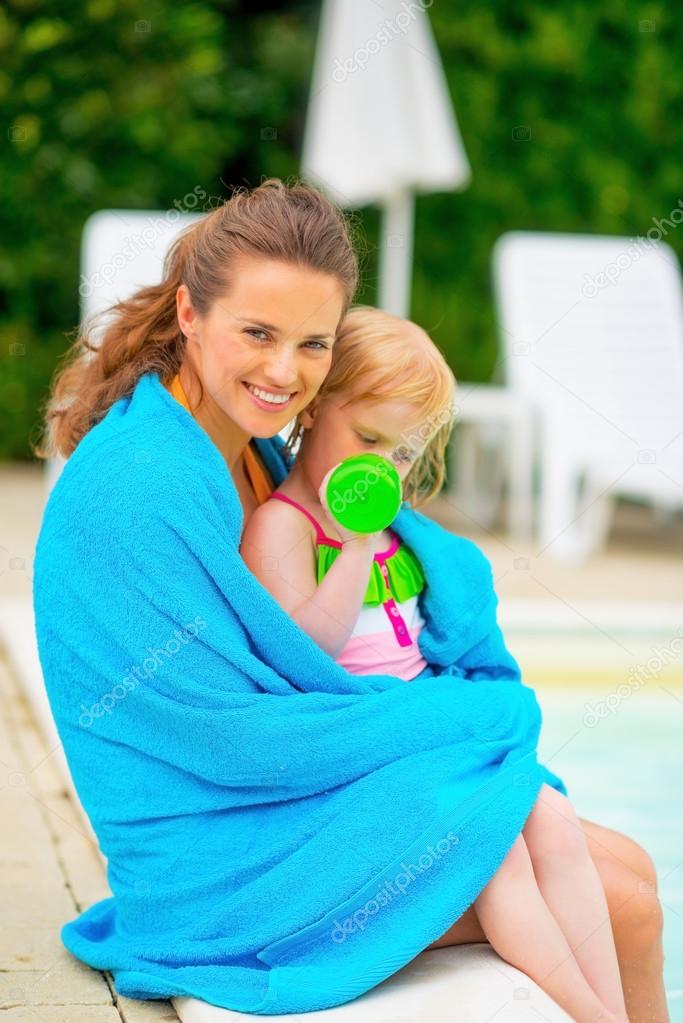 Happy mother and baby girl wrapped in towel sitting at poolside