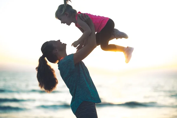 Junge Mutter wirft Kind bei Sonnenuntergang am Strand in die Luft — Stockfoto