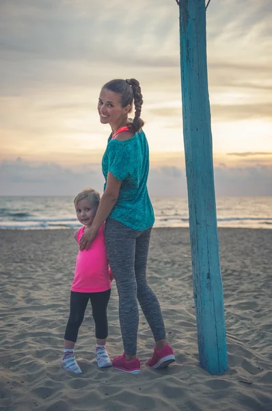 En glad ung mor på stranden står med sin datter - Stock-foto