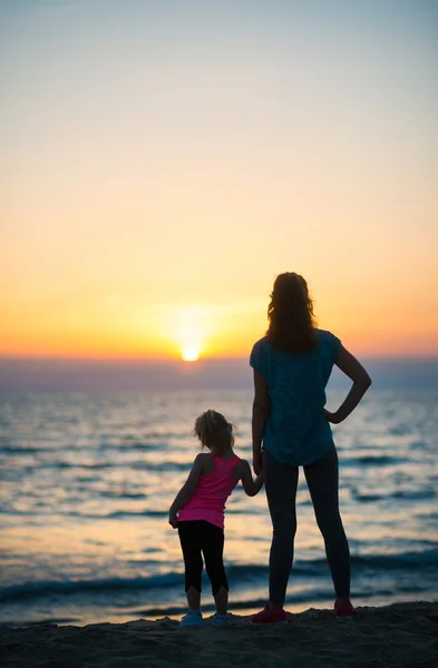Мать держит дочь за руку на закате, глядя на море — стоковое фото