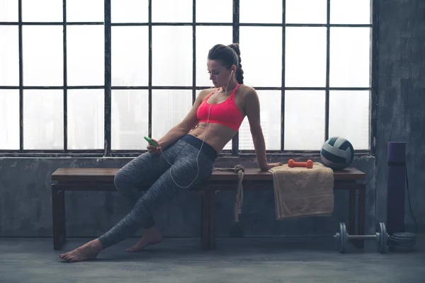 Fit mujer en el gimnasio loft sentado en el banco seleccionando música — Foto de Stock