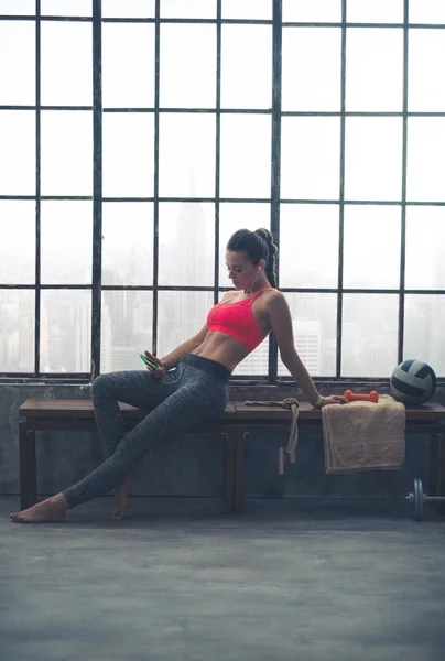 Woman reclining on bench selecting music in loft gym — Stock Photo, Image