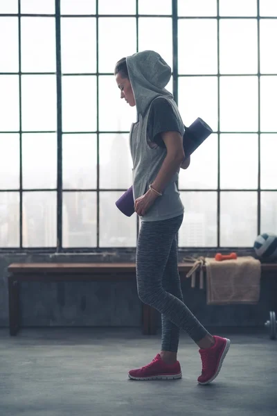 Fit woman wearing hoodie, carrying yoga mat in loft gym — 스톡 사진