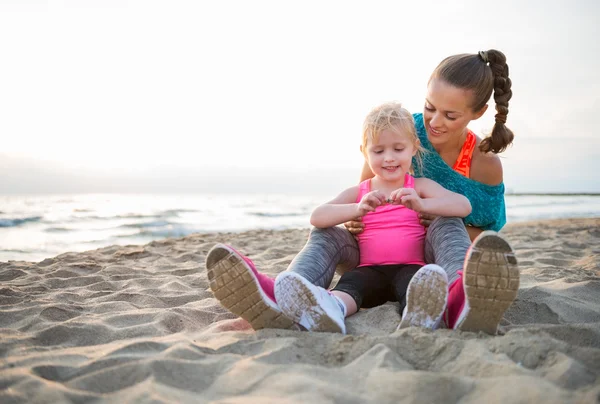 Ajuste joven madre e hija sentado en la playa al atardecer — Foto de Stock