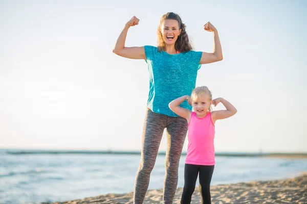 Glædelig mor og datter i fitness gear på strand bøjende arme - Stock-foto