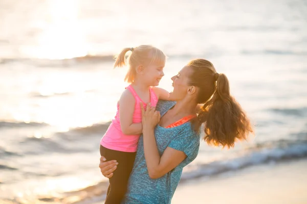 Happy mother holding daughter in her arms by water on beach — Stock Photo, Image